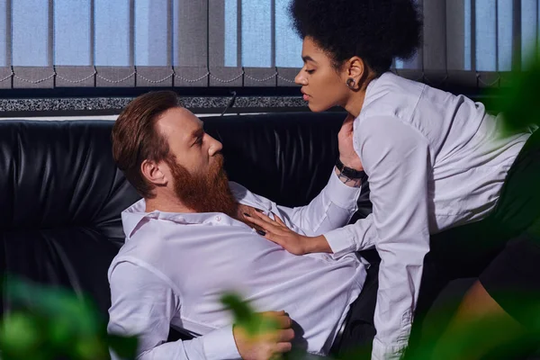 Sexy african american businesswoman seducing bearded colleague on couch in night office, side view — Stock Photo