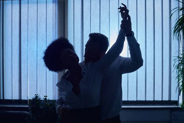 Dark silhouette of businessman holding hand and embracing african american woman in night office — Stock Photo