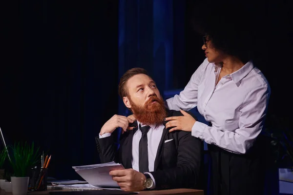 Спокуслива афроамериканська жінка обіймає плечі бородатого бізнесмена, який працює пізно в офісі — стокове фото