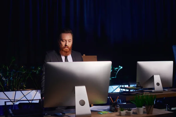 Hombre de negocios barbudo serio mirando el monitor de la computadora mientras trabaja hasta tarde en la oficina, turno de noche - foto de stock