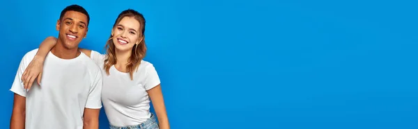 Feliz joven mujer abrazando amigo afroamericano en telón de fondo azul, diversidad cultural, bandera - foto de stock