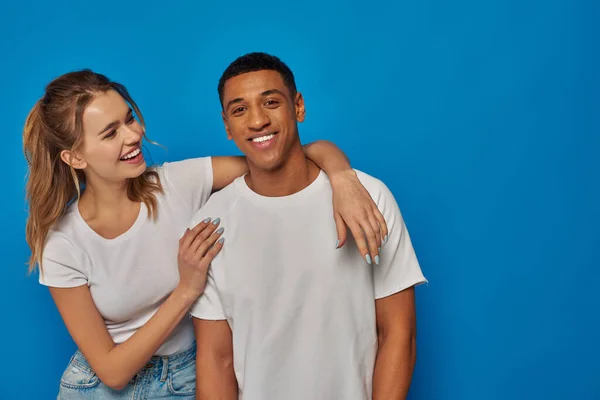 Feliz joven mujer abrazando afro-americano amigo en azul telón de fondo, emoción positiva y diversidad - foto de stock
