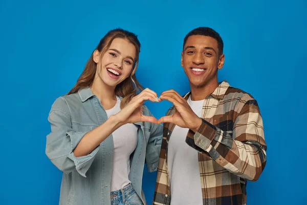 Alegre pareja interracial mostrando el signo del corazón con las manos y mirando a la cámara sobre fondo azul - foto de stock