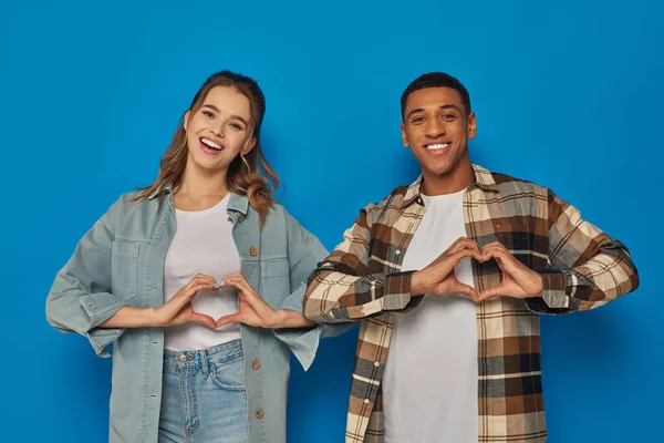Alegre pareja multicultural mostrando el signo del corazón con las manos y mirando a la cámara en el fondo azul - foto de stock