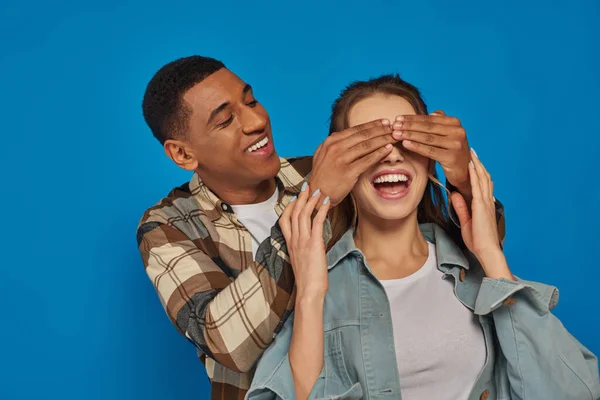 Hombre afroamericano excitado cubriendo los ojos de la mujer con la boca abierta sobre fondo azul, peekaboo - foto de stock