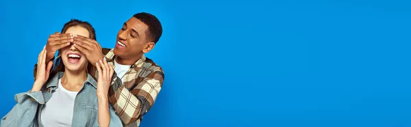 Excited african american man covering eyes of woman with open mouth on blue background, banner — Stock Photo