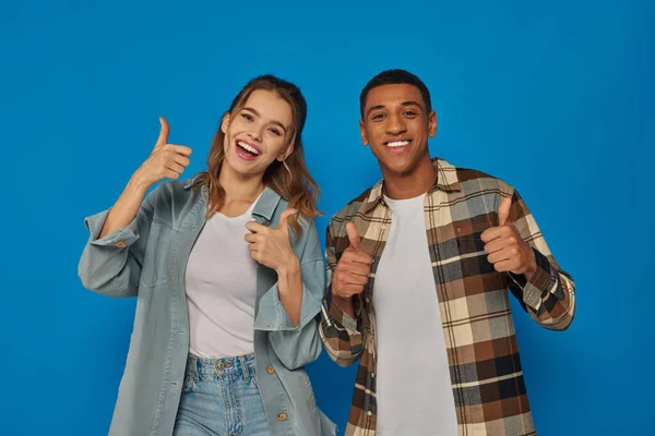 Glückliche interrassische Paar zeigt wie und Blick in die Kamera auf blauem Hintergrund, emotionale Reaktion — Stockfoto