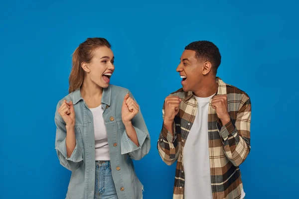 Feliz interracial pareja gritando de alegría mirando el uno al otro en azul telón de fondo, personas emocionales — Stock Photo