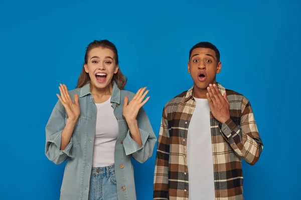 Feliz interracial pareja gritando de alegría mirando a la cámara en azul telón de fondo, reaccionando emocionalmente — Stock Photo