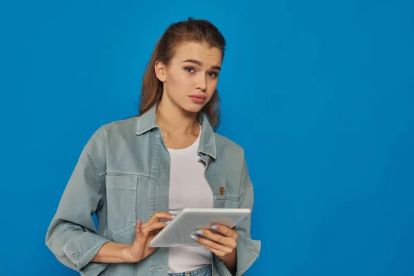 Hermosa mujer joven sosteniendo tableta digital y mirando a la cámara en el fondo azul, cara confusa - foto de stock