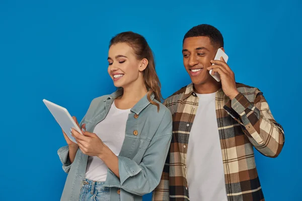 Счастливая межрасовая пара с помощью гаджетов на синем фоне, африканский американец с телефонным звонком — стоковое фото