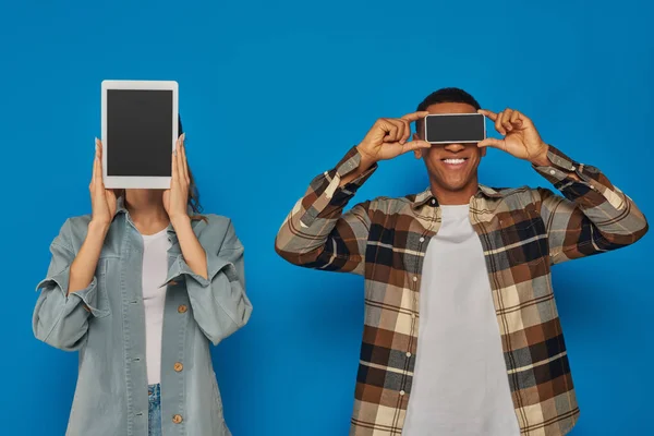 Interracial couple holding smartphone and digital tablet with blank screen on blue backdrop — Stock Photo