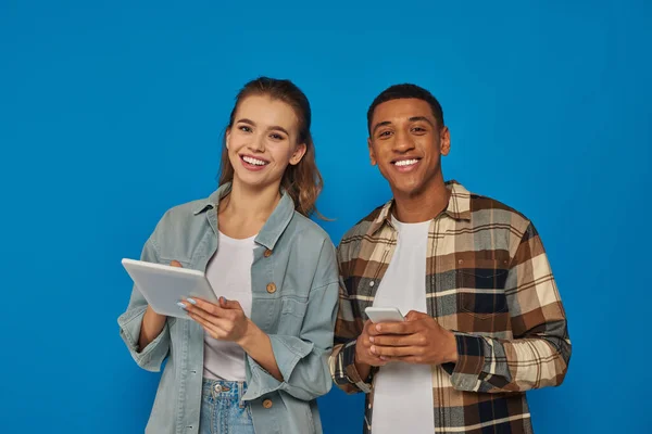 Feliz casal multiétnico segurando smartphone e tablet digital no fundo azul, olhar para a câmera — Fotografia de Stock