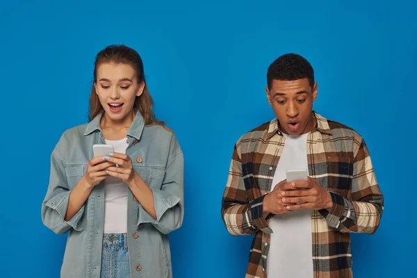Aufgeregter afrikanisch-amerikanischer Mann und staunende junge Frau chatten auf Smartphones vor blauem Hintergrund — Stockfoto