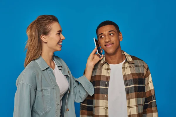 Glückliche Frau hält Smartphone in der Nähe von Ohr von afrikanisch-amerikanischen Kerl isoliert auf blauem Hintergrund — Stockfoto