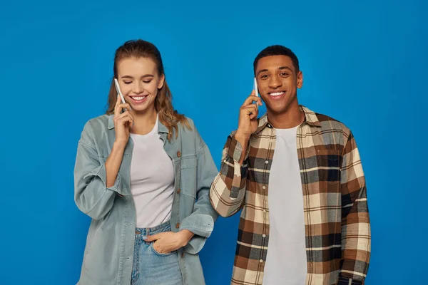 Cheerful woman and happy african american man having phone call, using smartphones on blue backdrop — Stock Photo