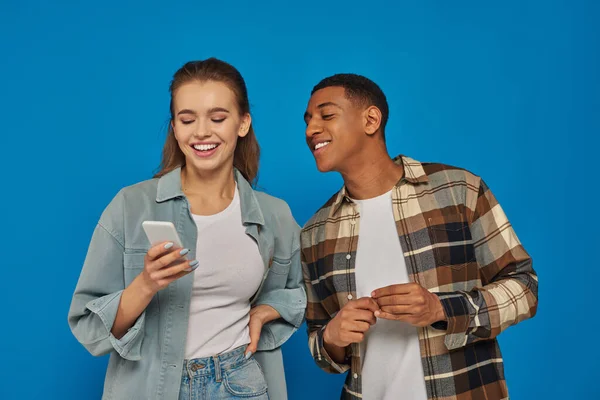 Happy african american man looking at  smartphone of female friend and smiling on blue backdrop — Stock Photo