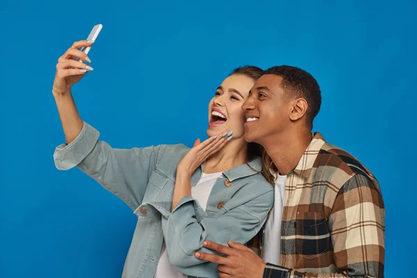 Alegre pareja interracial tomando selfie y sonriendo mientras mira el teléfono inteligente en azul telón de fondo - foto de stock