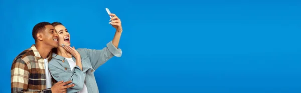 Cheerful interracial couple taking selfie while looking at smartphone on blue backdrop, banner — Stock Photo