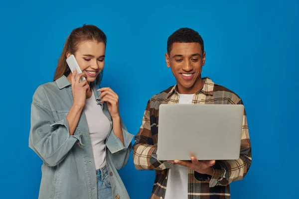 Freelancer afro-americano feliz usando laptop perto de mulher feliz falando no smartphone, fundo azul — Fotografia de Stock