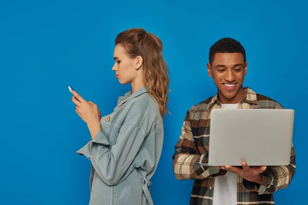 Happy african american freelancer using laptop near sad woman chatting on smartphone, blue backdrop — Stock Photo