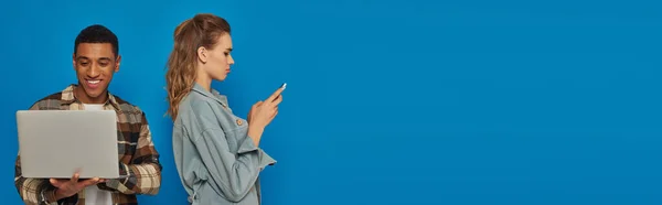 Freelancer afroamericano feliz usando el ordenador portátil cerca de mujer molesta charlando en el teléfono inteligente, bandera - foto de stock