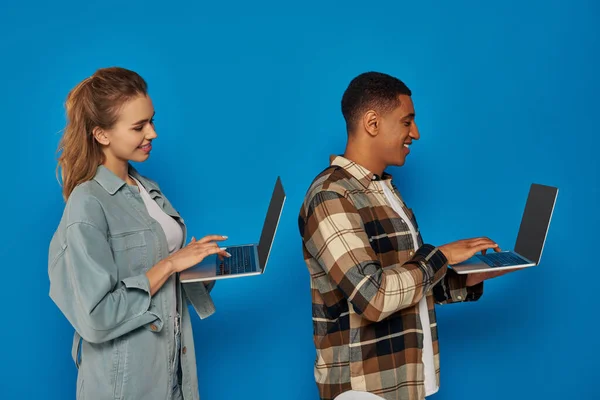Happy interracial freelancers using laptops on blue backdrop, diverse cultures man and woman, side — Stock Photo