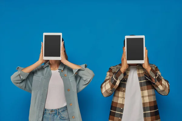 Межрасовые мужчина и женщина скрывая лицо с цифровыми таблетками на синем фоне, пустой сенсорный экран — стоковое фото