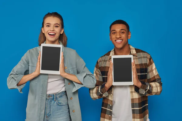 Happy interracial man and woman holding digital tablets with blank screen on blue backdrop — Stock Photo