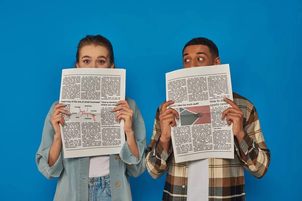 Межрасовые мужчина и женщина скрывают лица с газетами на синем фоне, читая новости — стоковое фото