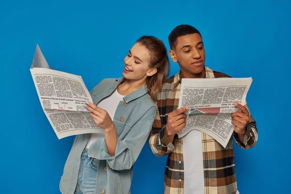 Hombre afroamericano feliz y mujer caucásica bonita leyendo noticias sobre telón de fondo azul, periódicos - foto de stock