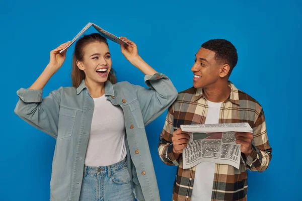 Happy woman covering head with newspaper near smiling african american man on blue backdrop — Stock Photo