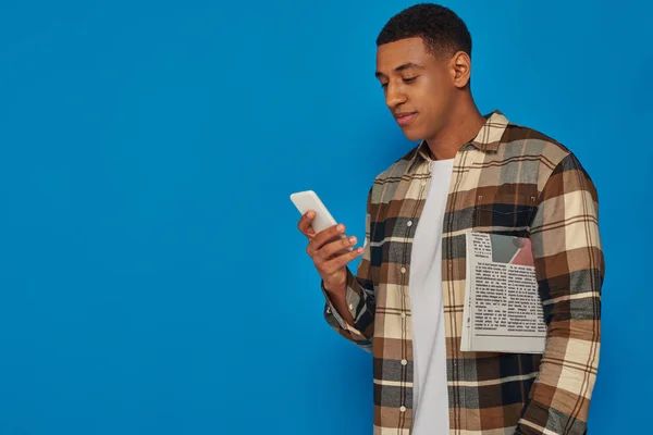 Hombre afroamericano en camisa a cuadros usando teléfono inteligente y sosteniendo periódico sobre fondo azul - foto de stock
