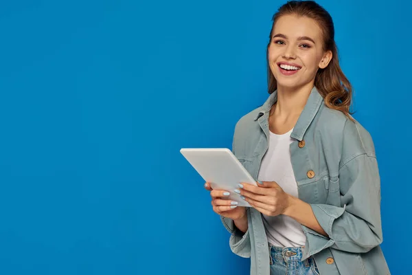 Glückliche junge Frau hält digitales Tablet in der Hand und lächelt in die Kamera auf blauem Hintergrund, soziale Medien — Stockfoto