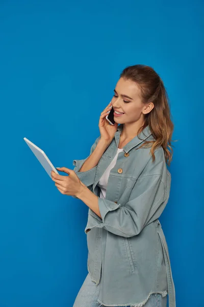 Счастливая молодая женщина говорит на смартфоне и с помощью цифрового планшета на синем фоне, подключение — стоковое фото
