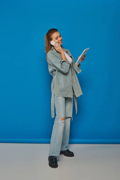 Joyeuse jeune femme parlant sur smartphone et utilisant une tablette numérique sur fond bleu, connectivité — Photo de stock