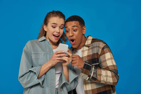 Stupito coppia interrazziale con la bocca aperta guardando smartphone su sfondo blu, utenti di social media — Foto stock