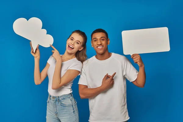 Amazed interracial couple pointing at blank placards on blue backdrop, thought and speech bubbles — Stock Photo