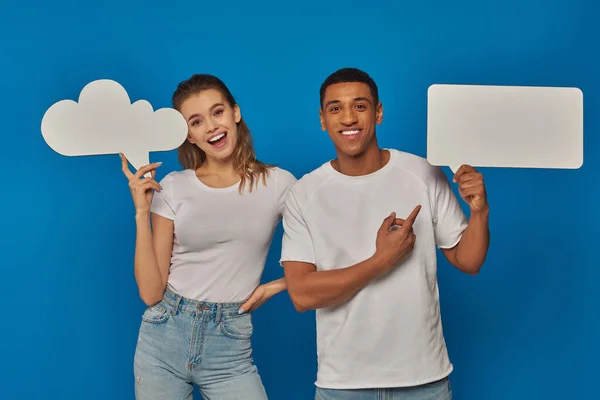 Casal interracial animado apontando para cartazes em branco no fundo azul, pensamento e bolhas de fala — Fotografia de Stock