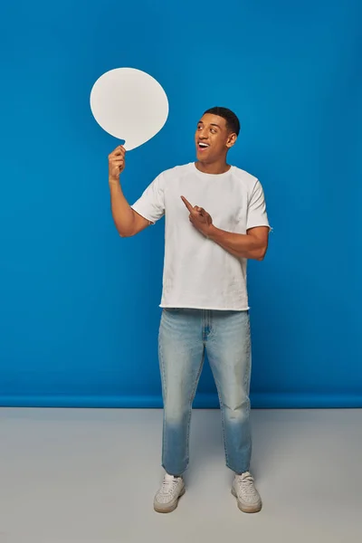 Схвильований афроамериканський чоловік у джинсах, що вказує на бульбашку мовлення на синьому фоні, повна довжина — стокове фото