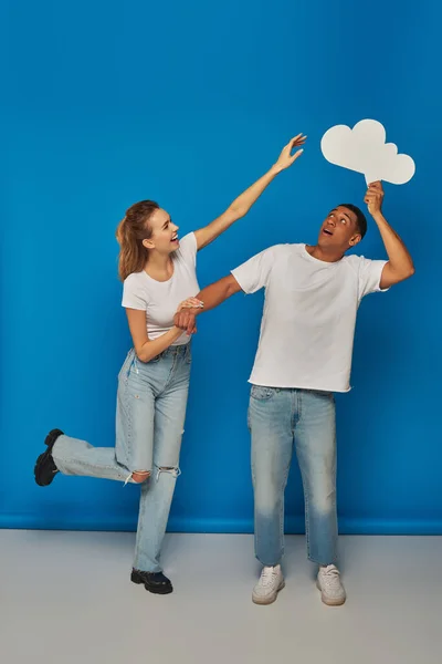 Woman with outstretched hand reaching thought bubble near african american man on blue backdrop, fun — Stock Photo