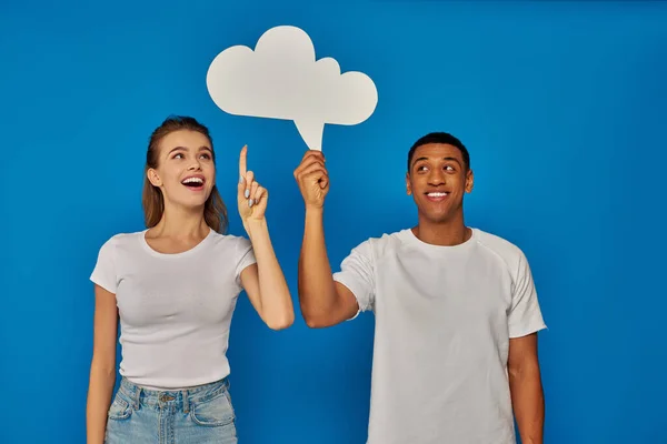 Heureux couple multiculturel souriant et pointant avec les doigts à bulle de pensée sur fond bleu — Photo de stock