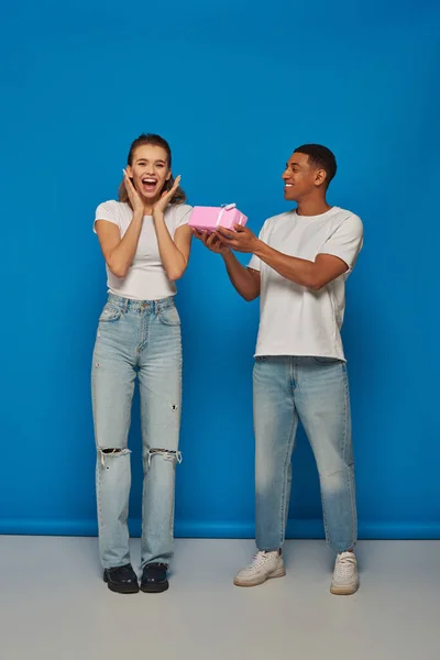 Happy african american man presenting wrapped gift to excited girlfriend on blue backdrop — Stock Photo