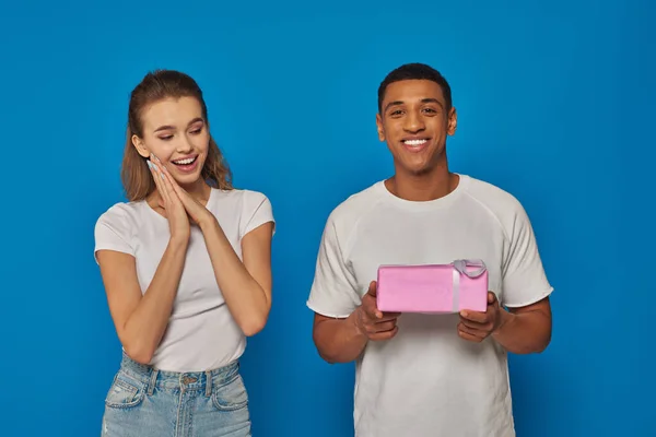 Joyeux homme afro-américain tenant cadeau enveloppé près de petite amie excitée sur fond bleu — Photo de stock