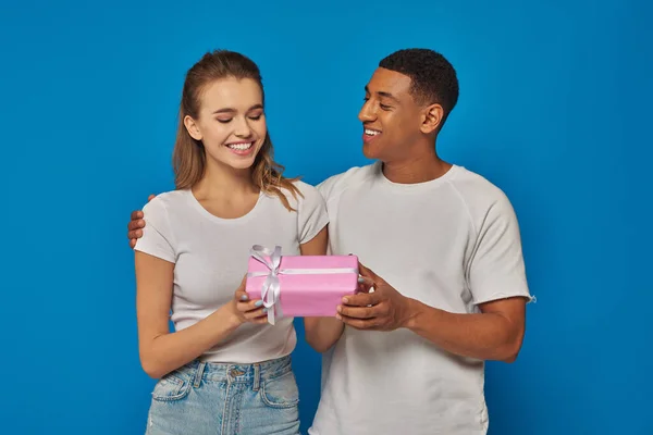 Freudiger afrikanisch-amerikanischer Mann überreicht verpacktes Geschenk an aufgeregte Freundin vor blauem Hintergrund — Stockfoto