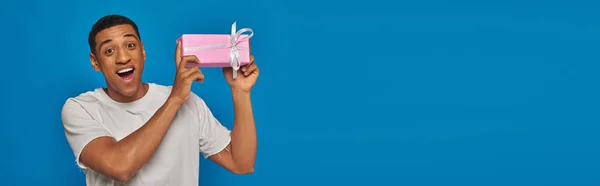Excited african american man holding wrapped present on blue background, festive occasions, banner — Stock Photo