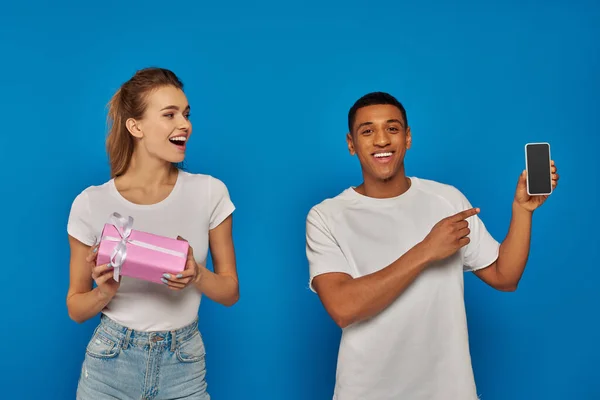 E-commerce, happy african american man pointing  at smartphone near woman with gift on blue backdrop — Stock Photo