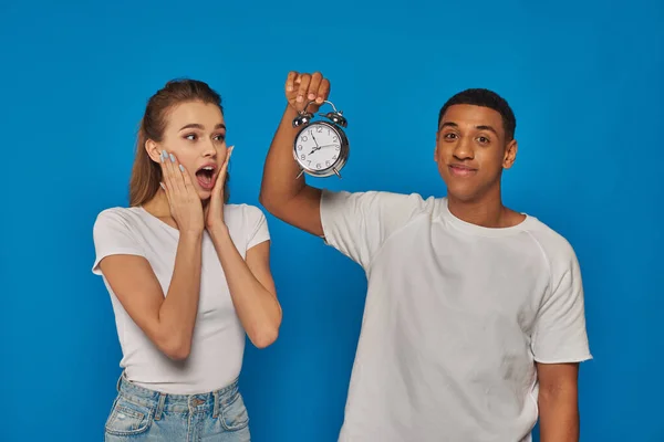 Morning time, happy african american man holding alarm clock near astonished woman on blue backdrop — Stock Photo