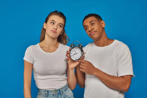 Hora de la mañana, dos pareja interracial disgustado celebración de reloj despertador retro en telón de fondo azul - foto de stock