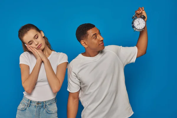 Morning time, african american man holding alarm clock near sleepy woman on blue background — Stock Photo